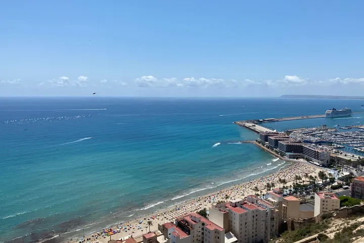 A long beach and a marina seen from the castle above, with turquoise water and a pier reaching out to sea.