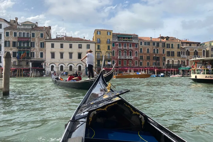 The bow of a gondola on the Grand Canal, with a gondolier and a row of palazzi ahead.