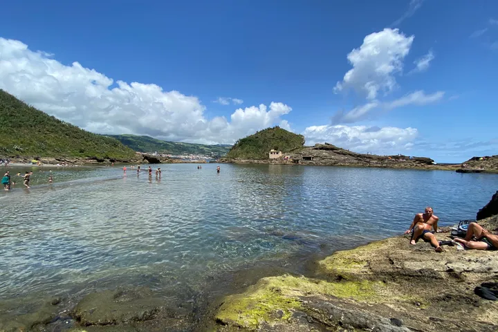 Shallow, clear water inside the ring of a volcanic islet, with people bathing and a blue sky.