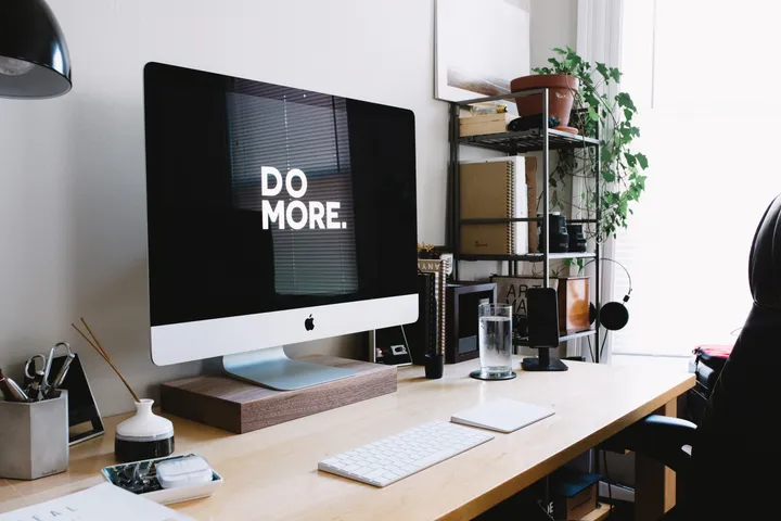 A desk with an iMac reading 'do more', a keyboard and a plant by the window.