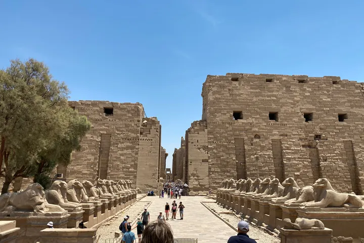 An avenue of ram-headed sphinxes leading to a stone pylon of Karnak temple, under a clear sky.
