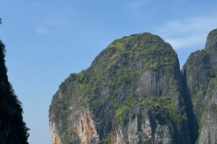 A limestone cliff covered in green, rising beside a second cliff under a blue sky.