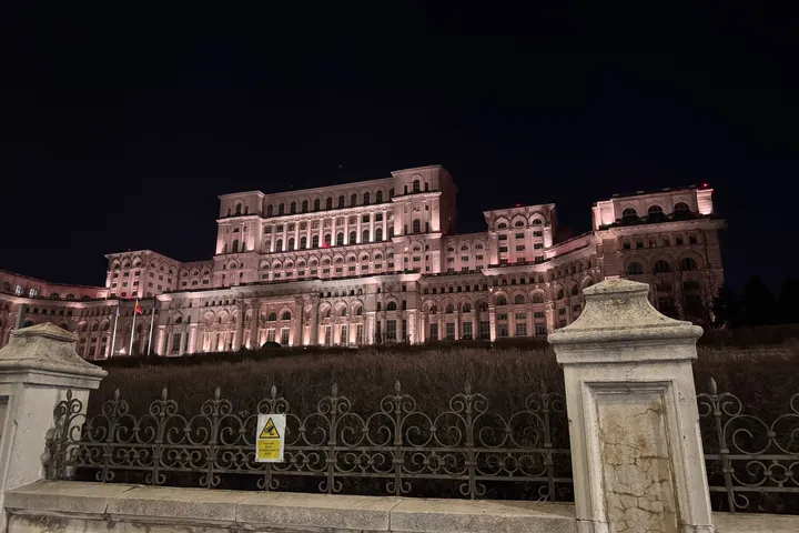 The Palace of the Parliament lit pink against a black night sky, behind an iron fence.