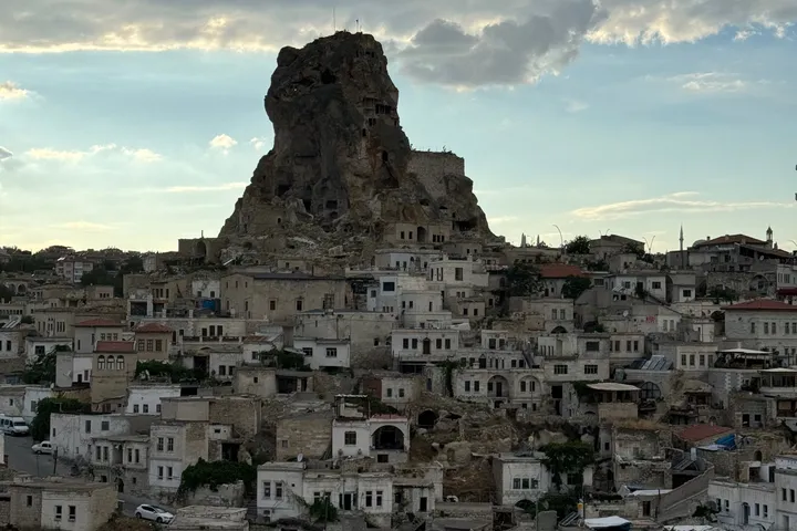 A rock outcrop rising above a town of pale stone houses under a partly cloudy sky.