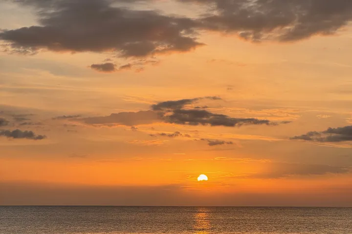The sun setting low over a calm sea, with orange light under a band of dark cloud.