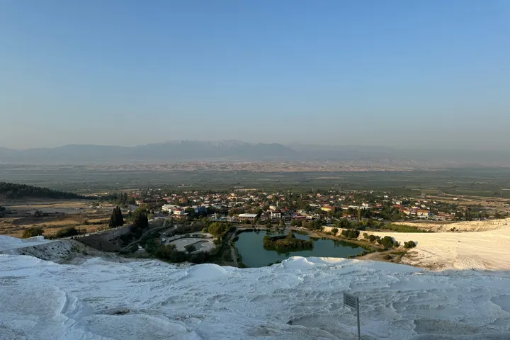 White travertine terraces above a small lake and a village, with a hazy plain and mountains beyond.